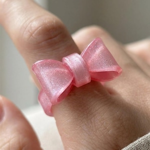 Può includere: Un anello rosa a forma di fiocco con dettagli glitterati. L'anello è realizzato in un materiale traslucido e si indossa su un dito. Il design a fiocco è una caratteristica importante, che aggiunge un tocco di fantasia al gioiello.