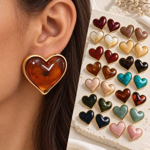 Può includere: Una collezione di orecchini a lobo a forma di cuore con una finitura liscia e lucida in vari colori, tra cui ambra, rosso, marrone, nero, verde acqua, verde e rosa. Ogni orecchino è incorniciato da un bordo in metallo color oro.