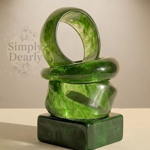 Può includere: Una pila di braccialetti verdi in resina e una base quadrata. I braccialetti sono di un verde traslucido con effetto marmorizzato. La base è verde scuro uniforme. Il testo "Simply Dearly" è in alto a sinistra.