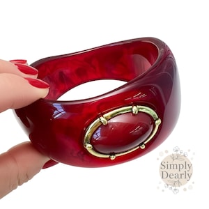 Può includere: Un braccialetto rosso a forma ovale con un elemento centrale ovale color oro. Il braccialetto ha un effetto marmorizzato ed è tenuto in mano. L'elemento centrale ha una pietra rossa.