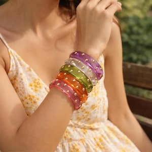 Può includere: Una collezione di braccialetti colorati e trasparenti adorna un polso. I braccialetti sono nelle tonalità di rosa, arancione, verde, trasparente e viola, ciascuno con piccoli accenti dorati. I braccialetti sono impilati, creando un accessorio vibrante ed elegante.