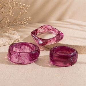 Può includere: Tre braccialetti in resina di forme e dimensioni diverse. I braccialetti sono di colore magenta traslucido con motivi a vortice. Uno è quadrato, gli altri due sono rotondi. I braccialetti sono esposti su una superficie neutra.