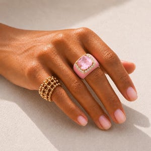 Può includere: Due anelli sono mostrati su una mano. Uno è dorato con un design a perline. L'altro è rosa con una pietra quadrata rosa chiaro e un bordo in oro e diamanti. Lo sfondo è di colore neutro.