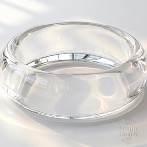 May include: A clear, circular glass bracelet with a thick band. The transparent material reflects light, creating highlights and shadows. The bracelet is a simple, elegant accessory.