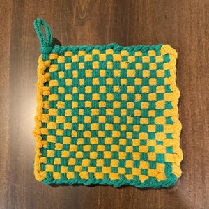 Op de afbeelding: Een vierkante, geweven pannenlap in een dambordpatroon van groen en geel. De randen zijn afgewerkt met geel garen en er is een groene lus bevestigd om op te hangen. De pannenlap is ontworpen voor gebruik in de keuken.
