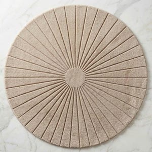 May include: A circular beige rug featuring a central solid circle with radiating lines extending to the outer edge. The rug has a textured, tufted surface with a consistent light tan color throughout, creating a sunburst pattern across the entire piece.
