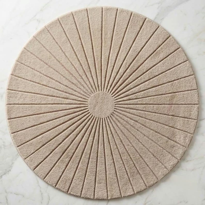 May include: A circular beige rug featuring a central solid circle with radiating lines extending to the outer edge. The rug has a textured, tufted surface with a consistent light tan color throughout, creating a sunburst pattern across the entire piece.