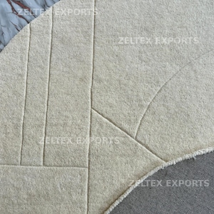 May include: A circular off-white rug featuring a textured surface with carved geometric lines. The rug has a thick, plush pile and a visible gray fabric backing along the curved edge. The text ZELTEX EXPORTS is printed across the surface.