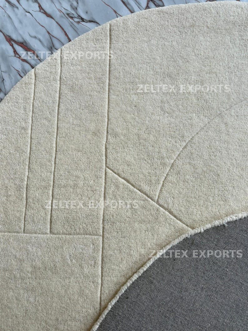 May include: A circular off-white rug featuring a textured surface with carved geometric lines. The rug has a thick, plush pile and a visible gray fabric backing along the curved edge. The text ZELTEX EXPORTS is printed across the surface.