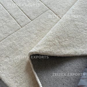 May include: A cream-colored area rug with a textured, tufted surface featuring a geometric pattern of straight lines. The rug is folded over to reveal a dark gray, woven fabric backing. The text ZELTEX EXPORTS is visible on the rug surface.