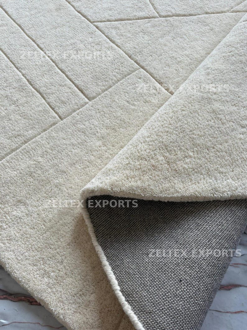 May include: A cream-colored area rug with a textured, tufted surface featuring a geometric pattern of straight lines. The rug is folded over to reveal a dark gray, woven fabric backing. The text ZELTEX EXPORTS is visible on the rug surface.