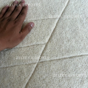 May include: A cream-colored area rug with a thick, plush, tufted texture featuring diagonal carved lines that create geometric sections across the surface. The text ZELTEX EXPORTS is printed in a light font across the rug's surface.