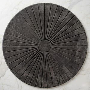 May include: A circular dark gray rug featuring a textured sunburst pattern. The design consists of a solid center circle with radiating lines extending outward to the edge, creating a symmetrical, fan-like appearance across the entire surface.