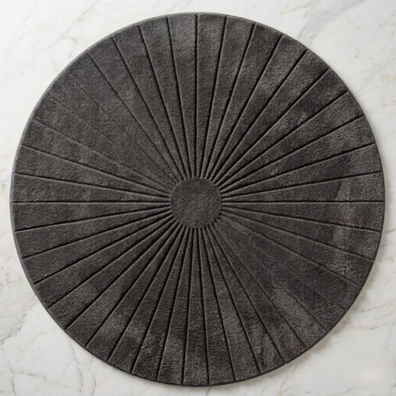 May include: A circular dark gray rug featuring a textured sunburst pattern. The design consists of a solid center circle with radiating lines extending outward to the edge, creating a symmetrical, fan-like appearance across the entire surface.