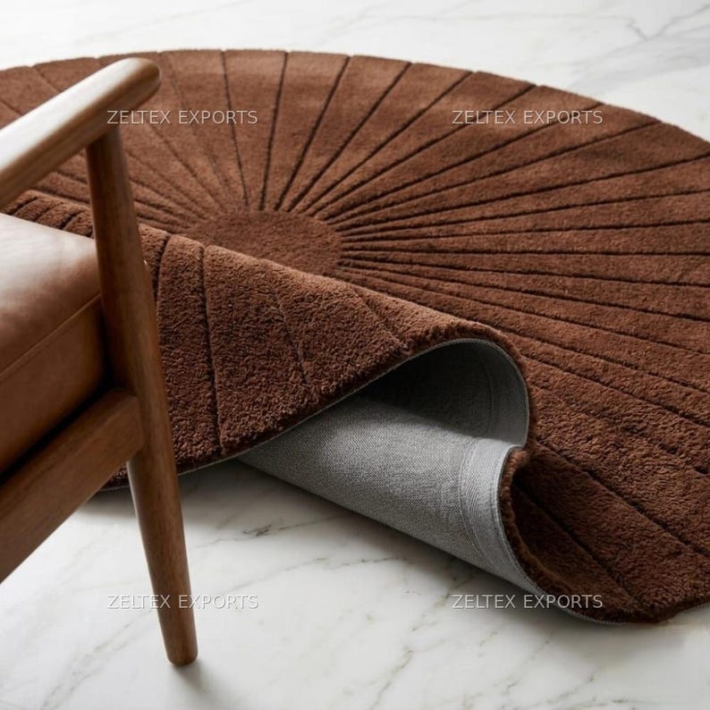 May include: A circular brown rug with a tufted texture and a radial sunburst pattern of carved lines radiating from the center. The rug is partially folded to reveal a smooth gray backing, and the text ZELTEX EXPORTS is visible.