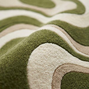 K&ouml;nnte beinhalten: Ein getufteter Teppich mit einem wellenf&ouml;rmigen, organischen Muster in Olivgr&uuml;n, Creme und Beige. Die Oberfl&auml;che weist eine dichte, flauschige Florstruktur mit klar definierten, geschwungenen Linien auf, die ein geschichtetes, r&auml;umliches Erscheinungsbild erzeugen.