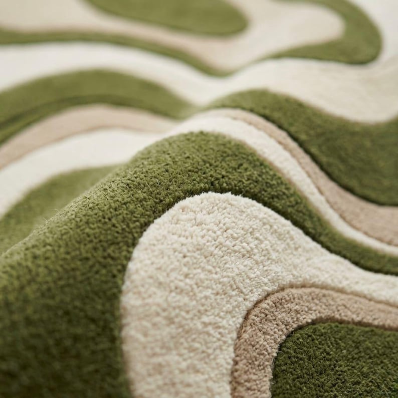 K&ouml;nnte beinhalten: Ein getufteter Teppich mit einem wellenf&ouml;rmigen, organischen Muster in Olivgr&uuml;n, Creme und Beige. Die Oberfl&auml;che weist eine dichte, flauschige Florstruktur mit klar definierten, geschwungenen Linien auf, die ein geschichtetes, r&auml;umliches Erscheinungsbild erzeugen.