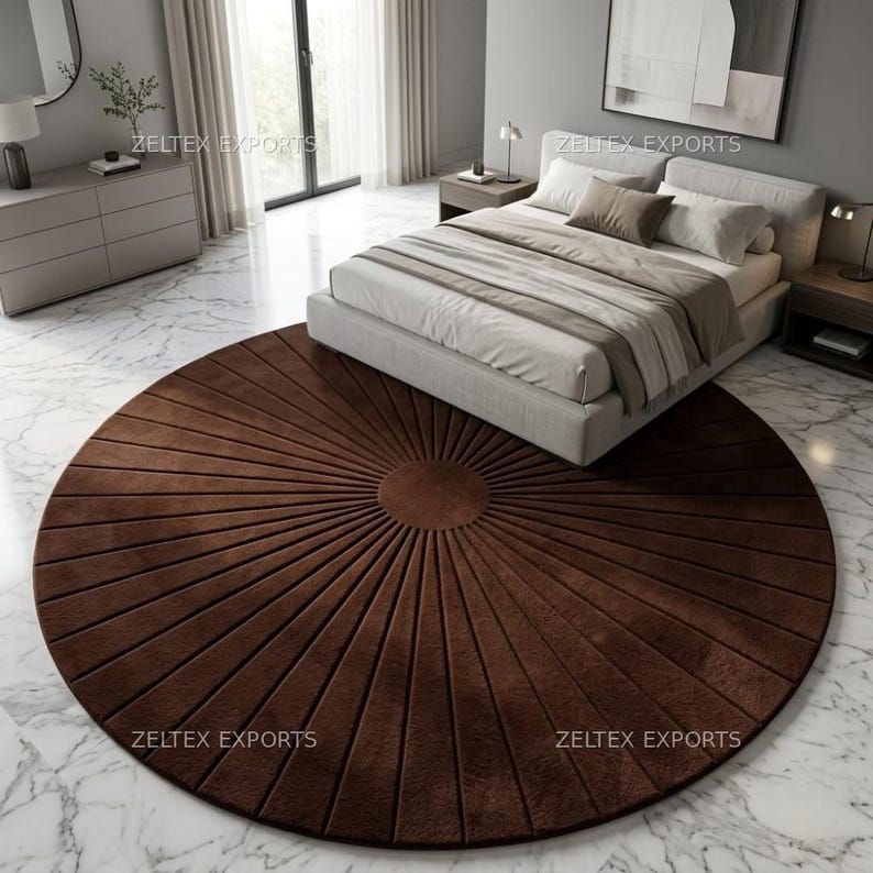 May include: A large, circular, dark brown area rug featuring a sunburst pattern with radiating lines extending from a solid center circle. The rug is placed on a white marble floor, partially positioned underneath a bed with light-colored bedding.