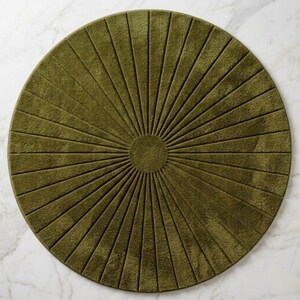 May include: A circular olive green rug featuring a central solid circle with radiating lines extending to the outer edge. The rug has a textured, plush pile surface with deep grooves creating a sunburst pattern across the entire round shape.