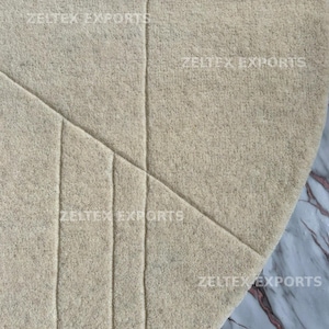 May include: A circular beige rug with a textured, felt-like surface featuring carved geometric lines. The text ZELTEX EXPORTS is repeated across the surface of the rug in a light font. The rug has a clean, rounded edge and a solid neutral color.