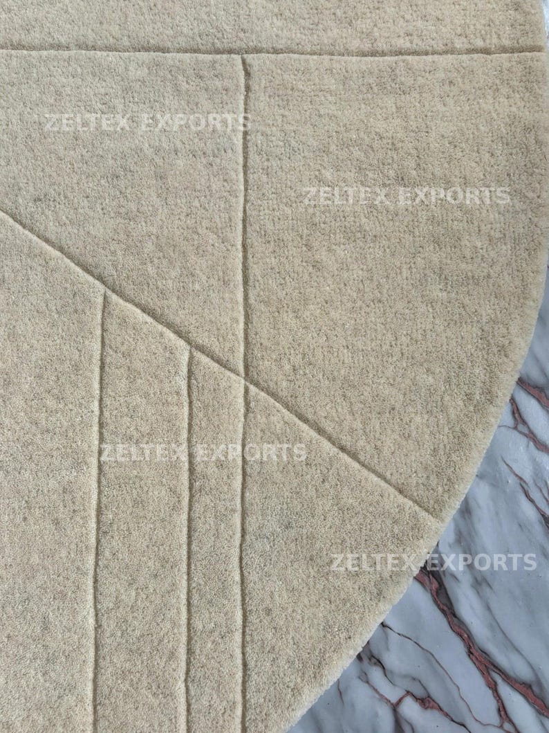 May include: A circular beige rug with a textured, felt-like surface featuring carved geometric lines. The text ZELTEX EXPORTS is repeated across the surface of the rug in a light font. The rug has a clean, rounded edge and a solid neutral color.