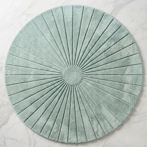 May include: A circular, sage green bath mat featuring a textured, sunburst pattern. The design consists of a solid central circle with radiating lines extending outward to the edge, creating a symmetrical, fan-like appearance across the surface.