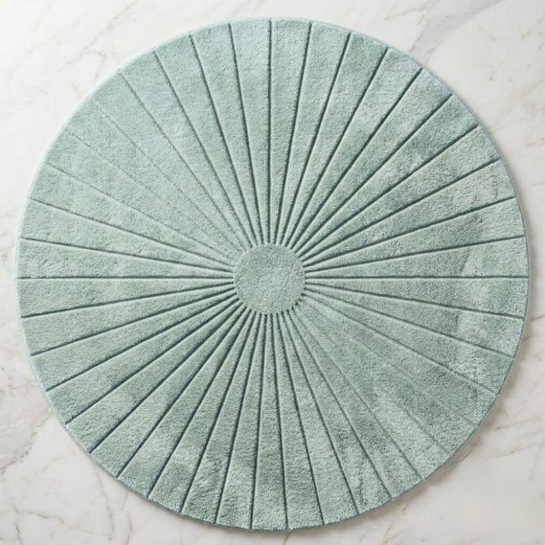 May include: A circular, sage green bath mat featuring a textured, sunburst pattern. The design consists of a solid central circle with radiating lines extending outward to the edge, creating a symmetrical, fan-like appearance across the surface.