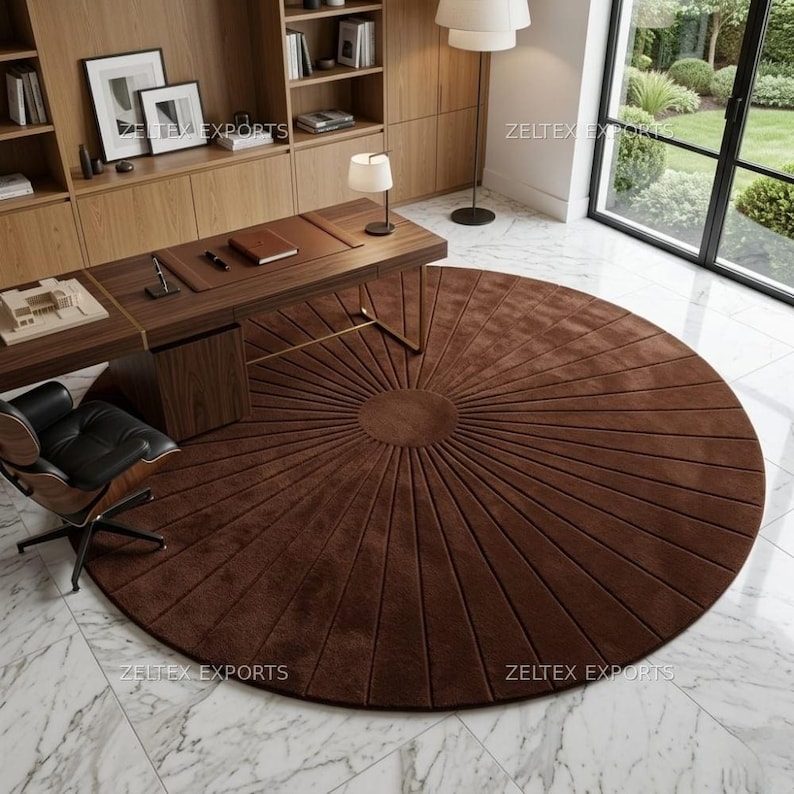 May include: A large, circular, dark brown rug featuring a sunburst pattern with radiating lines extending from a central solid circle. The rug is placed on a white marble floor in a room with a wooden desk, a black office chair, and shelving.