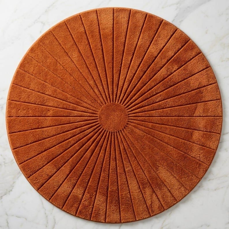 May include: A circular, rust-colored rug featuring a textured, sunburst-inspired pattern. The design consists of a solid center circle surrounded by radiating, recessed lines that extend outward to the edge of the rug, creating a uniform, symmetrical appearance.