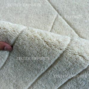 May include: A thick, off-white area rug with a dense, tufted pile texture. The surface features a subtle geometric pattern created by carved lines. The text ZELTEX EXPORTS is visible in multiple locations across the rug's surface.