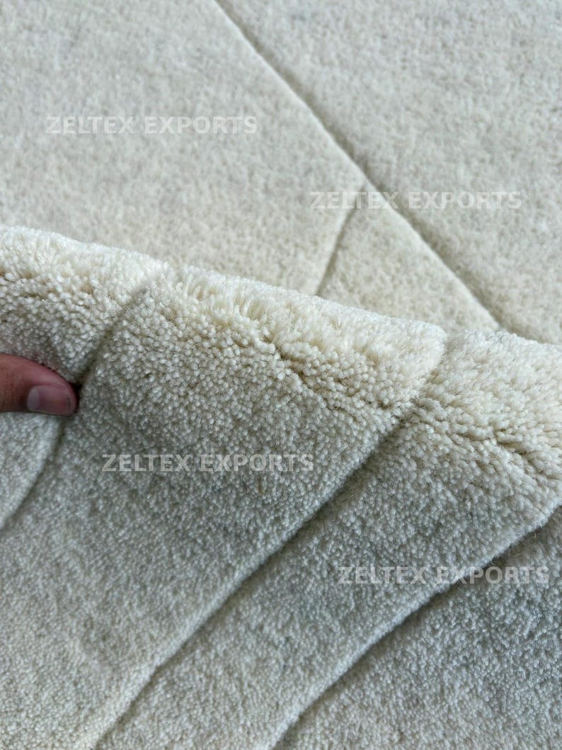 May include: A thick, off-white area rug with a dense, tufted pile texture. The surface features a subtle geometric pattern created by carved lines. The text ZELTEX EXPORTS is visible in multiple locations across the rug's surface.