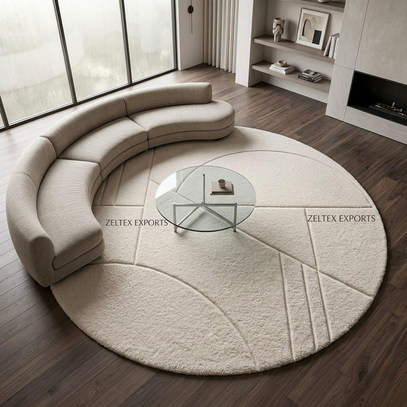 May include: A large, round, cream-colored area rug featuring a textured geometric pattern of lines and circles. The rug is positioned under a curved, beige sofa and a circular glass coffee table. The text ZELTEX EXPORTS is visible on the rug.