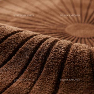 May include: A brown circular rug featuring a textured, tufted surface with a radial sunburst pattern of raised lines radiating from the center. The text ZELTEX EXPORTS is visible in four locations across the surface of the brown fabric material.