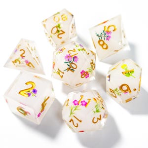 Puede incluir: Juego de siete dados de resina transparente con números dorados y diseños florales. Los dados presentan flores rosas, moradas y amarillas. Estos dados son para juegos de rol.