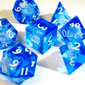 Puede incluir: Un conjunto de dados poliédricos azules translúcidos con vetas blancas y números blancos. Los dados tienen varias formas, incluyendo un cubo, una pirámide y un dado de 20 caras. Adecuado para juegos de mesa.