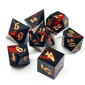 Puede incluir: Un conjunto de siete dados poliédricos para juegos de mesa. Los dados son negros con números dorados y acentos internos rojos y verdes. El conjunto incluye un D4, D6, D8, D10, D12, D20 y un dado de porcentaje.