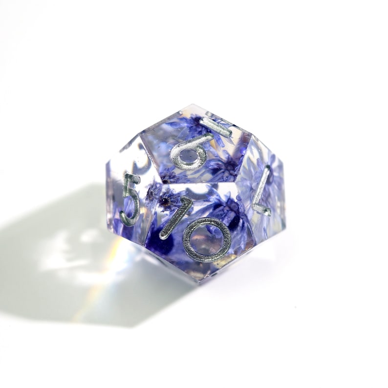 May include: A clear, multi-sided die with embedded blue flowers and silver numbering. The die is a D20, commonly used in tabletop role-playing games. The numbers are in a silver metallic font.