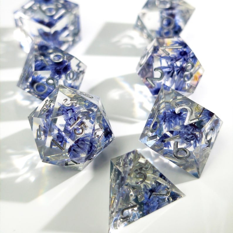 May include: A set of clear resin dice with blue flower inclusions and silver numbering. The dice are in various geometric shapes, including a d20, d12, and d6, and are arranged on a white surface. The dice are designed for tabletop gaming.