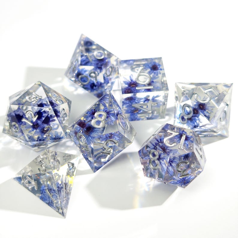 May include: Set of seven clear resin dice with blue flower inclusions and silver numbering. The dice are various shapes, including a d20, d12, d10, d8, d6, and d4, perfect for tabletop gaming.