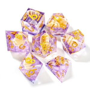 以下が含まれることがあります： 貝殻をテーマにした7つの多面体サイコロのセット。各サイコロは半透明の紫色で、中に貝殻が埋め込まれています。各面に金色の数字が見えます。テーブルゲームに最適です。