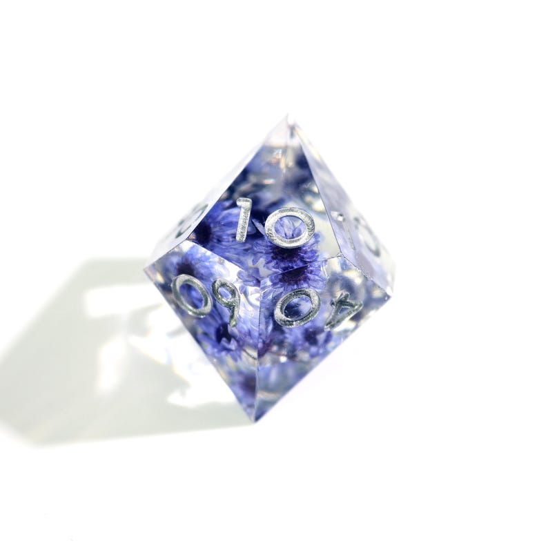 May include: A clear, multi-sided die with embedded blue flowers and silver numbering. The die is a unique gaming accessory, perfect for tabletop games or as a decorative item. The numbers are clearly visible.