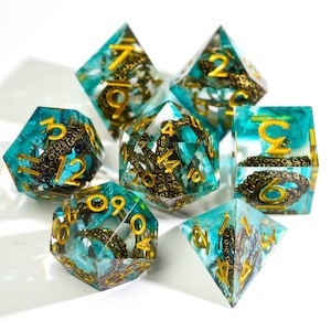 May include: A set of seven polyhedral dice with a teal and gold color scheme. Each die is transparent, with gold numbers and a bronze-colored octopus design inside. The dice are arranged on a white surface.