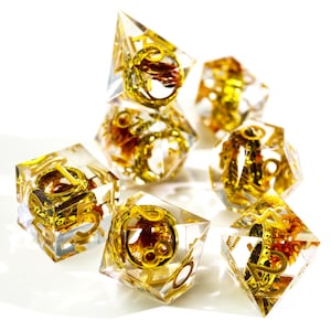 Puede incluir: Juego de siete dados de resina transparente con filigranas y numeración doradas. Los dados tienen varias formas poliédricas, incluyendo un d4, d6, d8, d10, d12, d20 y un dado de porcentaje. Para juegos de rol de mesa.