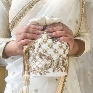May include: Cream-coloured drawstring pouch with detailed gold embroidery, sequins, and beadwork. The bag has a lattice design and floral accents. A gold tassel hangs from the side. The pouch is held by hands.