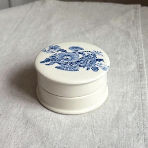 May include: A round, off-white ceramic trinket box with a lid. The top of the lid features a detailed blue floral pattern depicting a bouquet of flowers in a basket. The base of the box is smooth and cylindrical, matching the off-white color.