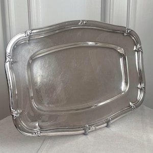 May include: A rectangular silver-toned serving tray with rounded corners and a raised, decorative border featuring subtle scrollwork details. The central surface is smooth and reflective, framed by a wide, contoured rim with a polished finish.