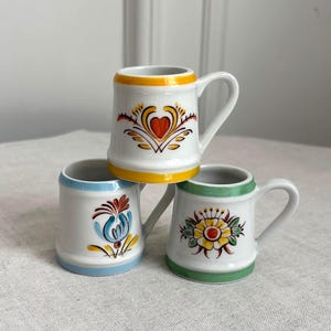Peut inclure: Trois tasses en céramique blanche avec anses, chacune présentant un motif peint à la main distinct. Une tasse a un bord jaune et un motif de cœur, une autre un bord bleu clair et une fleur, et la troisième un bord vert et une fleur.