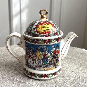 Op de afbeelding: Een witte keramische theepot met een feestelijke winterscène met mensen in een besneeuwd dorp en een verkoper van gepofte kastanjes. Het deksel is versierd met hulst, rode bessen, een goudkleurige knop en de tekst A Merry Christmas.