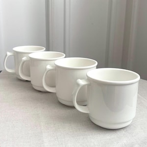 Könnte beinhalten: Ein Set aus vier schlichten, weißen Keramikbechern, die in einer diagonalen Linie angeordnet sind. Jeder Becher hat eine glatte, glänzende Oberfläche, einen abgerundeten Henkel an der Seite und einen leicht verjüngten Boden. Das Design ist einheitlich.