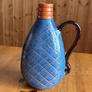 Puede incluir: Jarra de cerámica hecha a mano con cuerpo azul y cuello naranja. La superficie azul presenta un patrón de diamantes con líneas naranjas. El asa es de color marrón oscuro. Esta jarra decorativa es adecuada para servir bebidas.